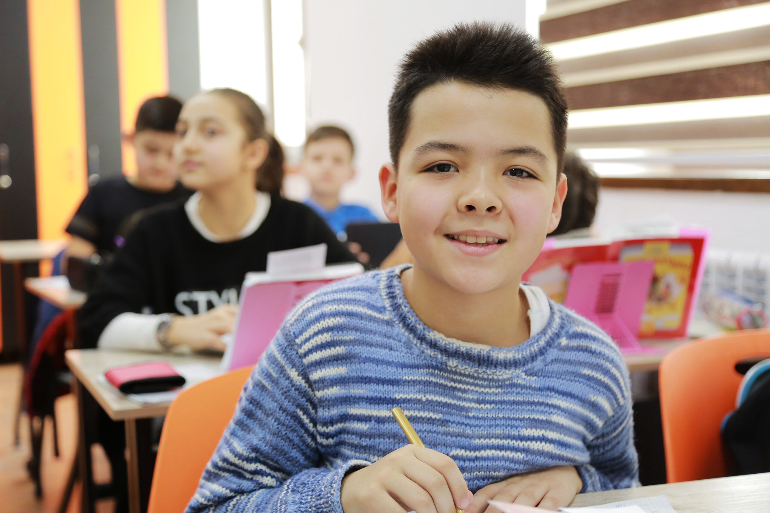 Ein Schüler schaut freundlich in die Kamera. Im Hintergrund beschäftigen sich weitere Schüler:innen mit ihrem Lernmaterial.